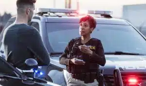 A policewoman taking a statement from a civilian outside her patrol car. The officer is a mature African-American woman in her 40s. She is talking with a young man in his 20s.