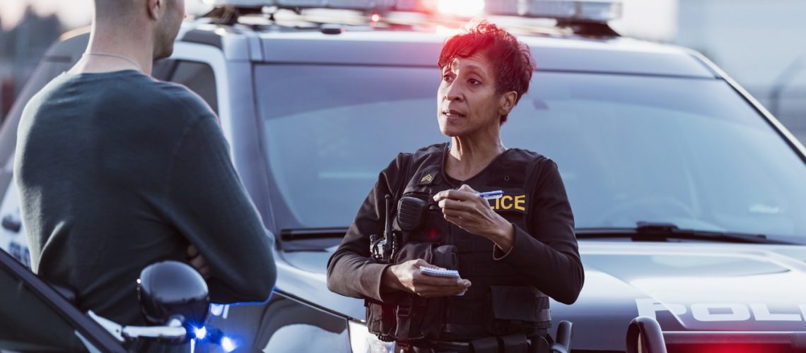 A policewoman taking a statement from a civilian outside her patrol car. The officer is a mature African-American woman in her 40s. She is talking with a young man in his 20s.